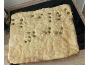 In der Größe eines Backblechs liegt ein helles Brot auf einer Arbeitsplatte. In den Teig sind Oliven eingelassen. Sie zeigen Linien und Ecken. Die Oliven als Punkte ergeben damit das Wort „Braille200“ in Brailleschrift.