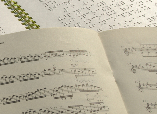Ein Notenheft in Schwarzschrift liegt auf einem Notenheft in Braille. Jemand hat mit einem Bleistift etwas darauf notiert.