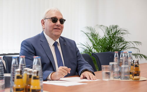 Hans-Werner Lange an einem Tisch, Unterlagen in Braille vor sich. Er hat graues Haar, trägt eine Sonnenbrille, Sakko, weißes Hemd und Krawatte.