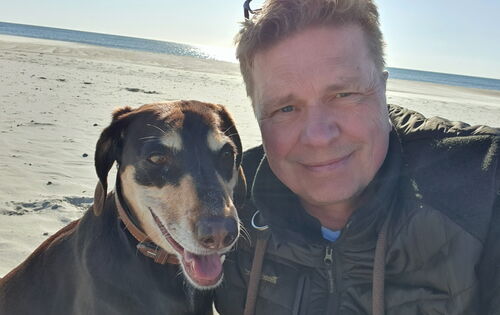 Ein Mann mit kurzen, hellen Haaren und einer dunklen Jacke macht ein Selfie mit einem braun-schwarzen Hund am Sandstrand. Beide schauen freundlich in die Kamera. Im Hintergrund sind Meer, blauer Himmel und Sonnenlicht zu sehen.