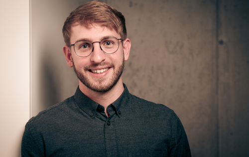 Fabian Rombach lächelt in die Kamera.  Er hat kurzes Haar, trägt eine Brille,  einen kurzen Bart und ein dunkles Hemd.