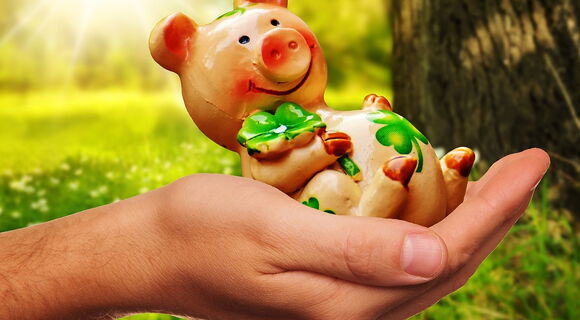 In einer menschlichen Hand liegt die Figur eines rosa Glücksschweinchens mit einem grünen Kleeblatt.