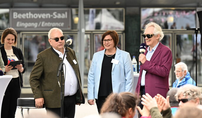 Auf der Außentribüne stehen Von links nach rechts Hans-Werner Lange (DBSV), Anne Reichmann (Nikolauspflege) und Arne Jöns (BSV Württemberg) und begrüßen die Festivalgäste.