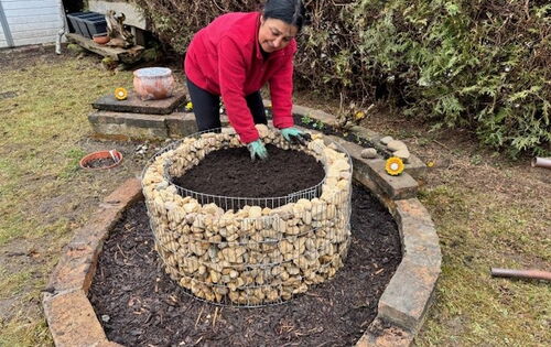 Selda beugt sich über ein steinernes Hochbeet, die Hände in der Erde. Sie lächelt und trägt eine leuchtend rote Jacke.