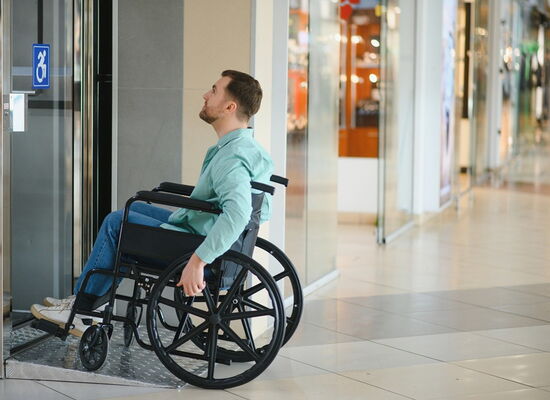 Ein Rollstuhlfahrer vor einer Tür in einem Einkaufszentrum. Auf der Glastür ist ein Aufkleber mit Rollstuhl angebracht.