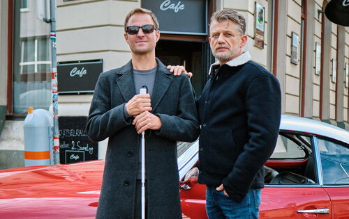 Das Foto zeigt Philipp Hochmair als Alex Haller mit Sonnenbrille und Langstock sowie Andreas Guenther als Niko Falk, der ihm eine Hand auf die Schulter legt.