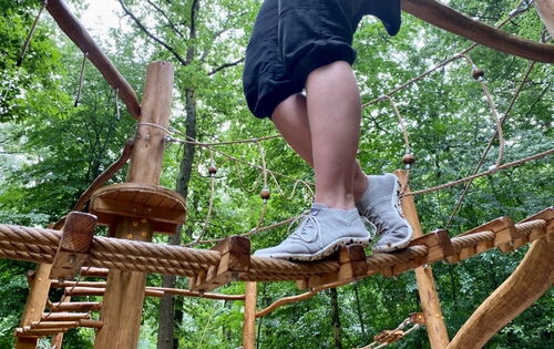 Frauenbeine in hellen geschnürten Barfußschuhen. Die Trägerin balanciert über ein dickes Seil in einem Hochseilgarten. Im Hintergrund stehen Bäume.
