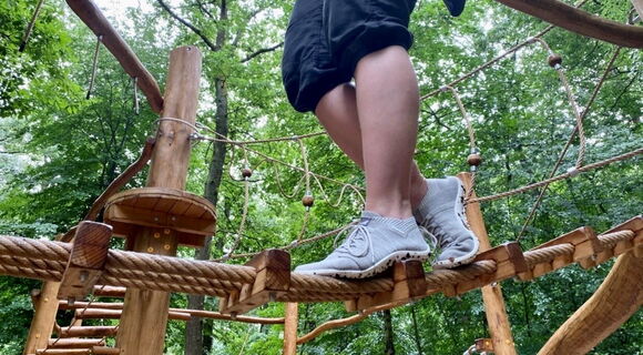 Frauenbeine in hellen geschnürten Barfußschuhen. Die Trägerin balanciert über ein dickes Seil in einem Hochseilgarten. Im Hintergrund stehen Bäume.