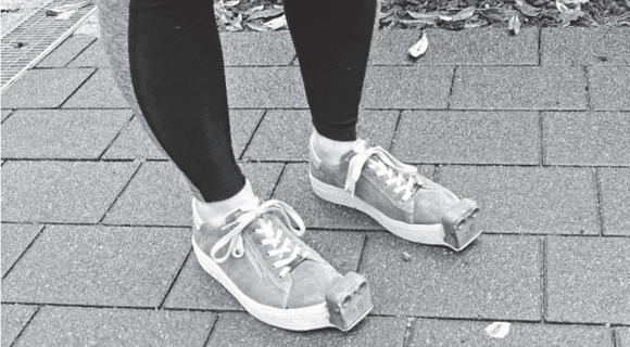 Ein Paar Beine in schwarzen Leggings und mit den besonderen Schuhen: An deren Spitze ist jeweils das InnoMake-Gerät befestigt, ein kleiner Kasten aus Metall.