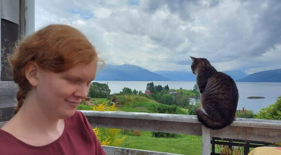Solveig Marie Oma steht links im Bild und blickt nach unten. Rechts sitzt eine getigerte Katze auf einem Geländer. Im Hintergrund sind ein Fjord, grüne Landschaft, Berge und ein bewölkter Himmel zu sehen.