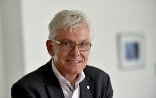 Ein älterer Mann mit weißem Haar, Brille und hellem Teint trägt ein dunkles Sakko über einem weißen Hemd. Er lächelt freundlich und blickt direkt in die Kamera. Der Hintergrund ist neutral und unscharf, mit einem gerahmten Bild an der Wand.