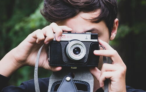 Ein junger Mensch mit dunklen Haaren hält eine analoge Kamera vor sein Gesicht und fotografiert. Die Kamera hat ein Vintage-Design mit schwarzem Gehäuse und silbernen Details. Der Hintergrund ist grün und unscharf.