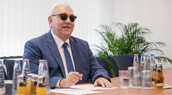 Hans-Werner Lange an einem Tisch, Unterlagen in Braille vor sich. Er hat graues Haar, trägt eine Sonnenbrille, Sakko, weißes Hemd und Krawatte.