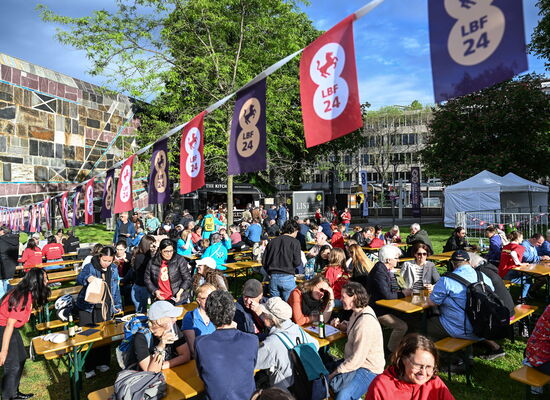 Berliner Platz neben der Liederhalle. Auf einer Rasenfläche stehen im Sonnenlicht Bierbänke und Tische, an denen zahlreiche Menschen sitzen. Über ihnen ist eine Leine gespannt, an der Fähnchen mit dem Logo des Louis Braille Festivals hängen.