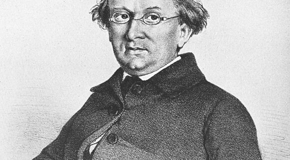 Zeichnung eines Mannes, Eduard Mörike, mit Brille und gewelltem Haar. Er trägt einen dunklen Mantel mit mehreren Knöpfen und sitzt mit leicht geneigtem Kopf. Der Hintergrund ist schlicht.