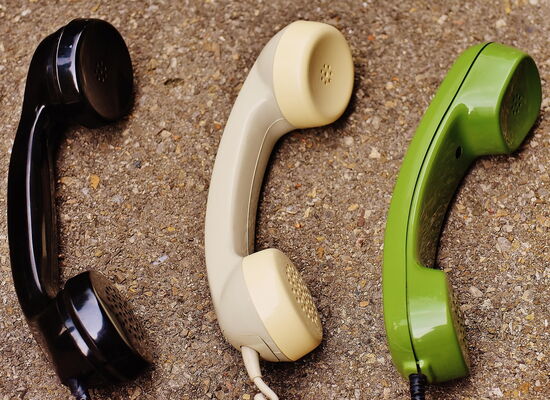 Drei klassische Telefonhörer in Schwarz, Beige und Grün liegen nebeneinander auf einem Korkuntergrund.