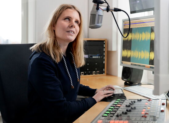 Nina Odenius in einem Studio am Mischpult und in ein auf Kopfhöhe angebrachtes Mikrofon sprechend. Sie hat langes blondes Haar und helle Augen.