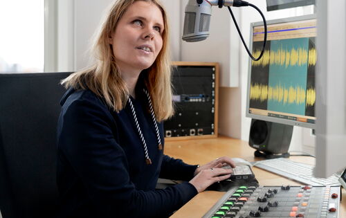Nina Odenius in einem Studio am Mischpult und in ein auf Kopfhöhe angebrachtes Mikrofon sprechend. Sie hat langes blondes Haar und helle Augen.