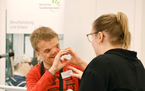 Ein Junge im orangen T-Shirt macht taktile Gebärden mit einer Frau mit blondem Zopf. Sie stehen vor einem Banner des Deutschen Taubblindenwerks. Taktile Gebärden sind eine Kommunikationsform von Menschen mit Taubblindheit.