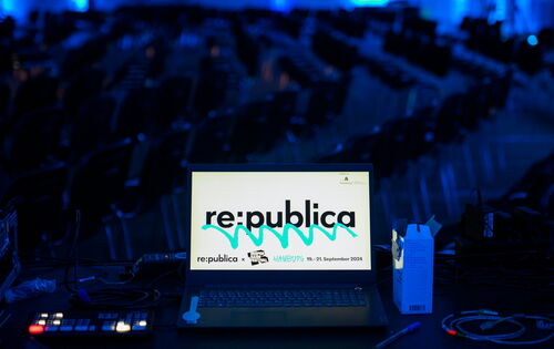 Ein Laptop auf einem Technik-Tisch zeigt das Logo der „re:publica". Im Hintergrund sind viele leere Stuhlreihen in einem blauen Licht beleuchteten Raum zu sehen. Kabel und Technikgeräte liegen auf dem Tisc