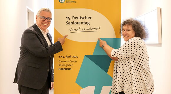 Ein Mann und eine Frau stehen neben einem Aufsteller vom 14. Deutschen Seniorentag und zeigen mit dem Finger darauf. Sie haben die Augen weit geöffnet und lachen. Er trägt einen dunklen Anzug, sie trägt eine gepunktete Bluse und hat lockige, graue Haare.