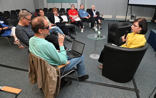 Workshop in einem größeren Raum: Die Teilnehmerinnen und Teilnehmer sitzen in einer Reihe; vorne befinden sich ein Mann und eine Frau, die jeweils ein Mikro in der Hand halten.