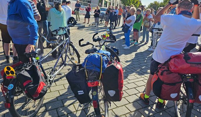 Die Tandems sind bereit: Sie sind mit Fahrradtaschen bepackt, einer der Fahrer setzt sich einen Helm auf. Rund 50 Menschen stehen im Halbkreis beisammen für die Verabschiedung.