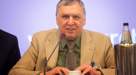 Wolfgang Angermann hat kurzes graues Haar. Er trägt ein helles Sakko und ein dunkles Hemd und spricht, an einem Tisch sitzend, in ein Mikrofon.