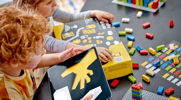 Zwei Kinder sitzen an einem Tisch. Vor sich liegt ein Karton mit Braille-Bricks, darum herum Legosteine.