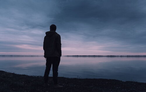 Die Silhouette einer männlich anmutenden Person, die alleine auf ein weites Gewässer blickt. Das Bild ist in dunklen Farben gehalten.
