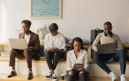 Mit unterschiedlichen Geräten am selben Projekt arbeiten: Vier junge Menschen sitzen nebeneinander auf einer breiten Bank und gucken jeweils in einen Laptop, ein Tablet oder ein Handy. Es sind vier junge Personen in schlichter, moderner Kleidung.