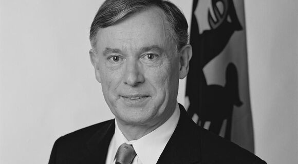 Horst Köhler schaut leicht lächelnd in die Kamera. Er hat sein Haar auf der linken Seite gescheitelt. Er trägt einen schwarzen Anzug und eine helle Krawatte. Im Hintergrund steht eine Fahne mit Reichsadler-Abbildung.