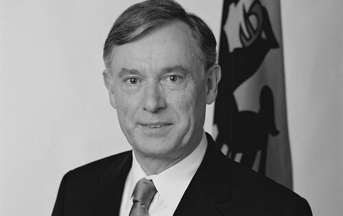 Horst Köhler schaut leicht lächelnd in die Kamera. Er hat sein Haar auf der linken Seite gescheitelt. Er trägt einen schwarzen Anzug und eine helle Krawatte. Im Hintergrund steht eine Fahne mit Reichsadler-Abbildung.
