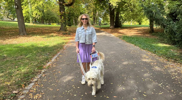 Lisa Mümmler geht mit Führhund Harry im Führgeschirr auf einem Weg im Park, das Ambiente ist herbstlich. Sie trägt Ray-Ban Meta Glasses, eine helle Jeansjacke und ein lilafarbenes Kleid.