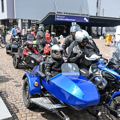 Mehrere Motorräder mit Beiwagen stehen vor der Liederhalle. Sie sind besetzt mit Menschen in schwarzer Motorradkleidung. Alle tragen schwarze Motorradhelme. Die Visiere sind hochgeklappt. Ganz vorn steht ein blau-schwarz lackiertes Motorrad.