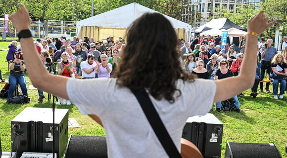 Das Bild ist von hinten über die Schulter einer Frau mit dunklen, langen Haaren in weißem T-Shirt aufgenommen. Sie wirft vor einem ausgelassenen Publikum motiviert die Arme in die Höhe.
