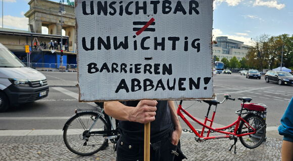 Ein Mann hält ein Schild in die Kamera: „UNSICHTBAR ≠ UNWICHTIG – BARRIEREN ABBAUEN“. Er steht vor dem Brandenburger Tor. Im Hintergrund ist ein rotes Tandem zu sehen.