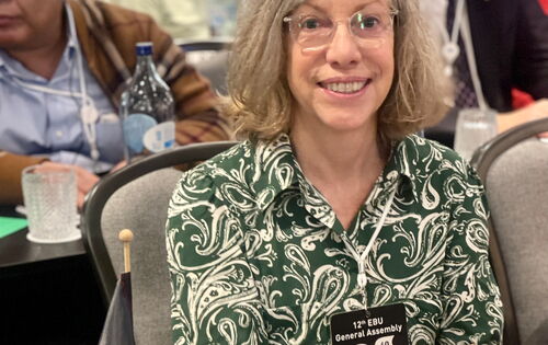 Sabine Ström sitzt während der Generalversammlung an einem Tisch mit Deutschland-Fähnchen und dem Schild „Germany“. Sie trägt mittellanges helles Haar, eine Brille und ein Kleid mit floralen Mustern. Hinter ihr sind weitere Teilnehmer zu erkennen.