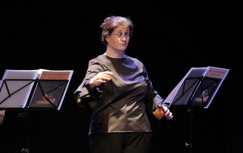 Bárbara Martín Muñoz steht auf einer Bühne, mit beiden Händen spielt sie je eine Kastagnette. Sie blickt auf Noten auf einem Notenständer. Ihr Haar hat sie hochgebunden, sie trägt eine Brille.