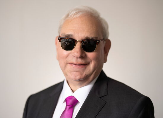 Portrait von Hans-Werner Lange. Er hat kurze, weiße Haare und ein volles Gesicht. Er trägt ein weißes Hemd, darüber ein dunkles Jackett, dazu eine purpurfarbene Krawatte und eine dunkel getönte Brille. Er schaut lächelnd in die Kamera.