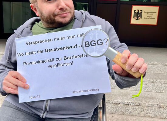 Ein Mann mit kurzen blonden Haaren hält eine Lupe und ein Schild in den Händen. Darauf steht: „Versprechen muss man halten. Wo bleibt der Gesetzentwurf zum BGG? Privatwirtschaft zur Barrierefreiheit verpflichten!“
