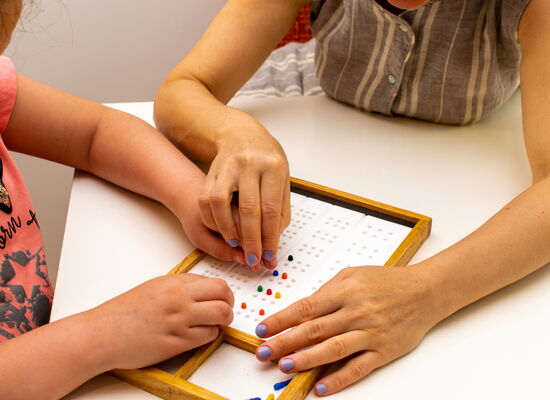 Die Hände eines Mädchens und einer Lehrerin an einem Braille-Board zum Lernen des Alphabets. Sie ertasten die Erhebungen. Beide tragen gemusterte Oberteile und haben lackierte Fingernägel.