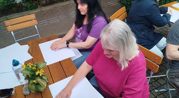 Zwei Frauen sitzen an einem Holztisch und studieren mit den Fingern je eine Wanderkarte in Braille. Die Frau links ist Yvonne Bühr. Sie hat langes dunkles Haar.