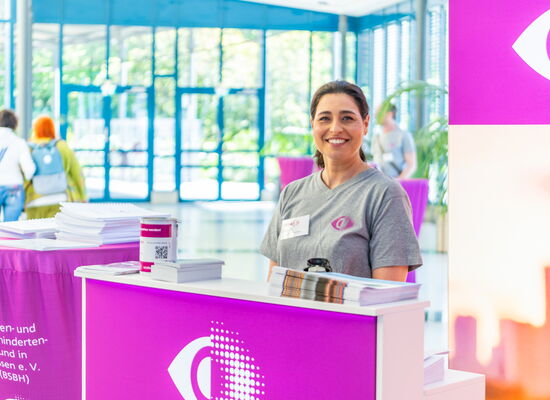 Bei der Jubiläumsfeier in Hessen: Eine Frau steht lächelnd an einem Infostand in einem Gebäude mit Glasfront. Das Rasterauge des DBSV prangt hinter ihr, am Tisch des Standes und auf ihrem T-Shirt.