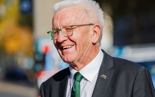 Winfried Kretschmann lachend an einem sonnigen Tag. Er trägt eine grüngerahmte Brille zur grünen Krawatte. Er ist ein Mann Ende 70 mir weißen Haaren.