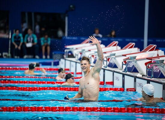 Taliso Engel winkt mit einem breiten Lachen aus dem Wasser in einem Schwimmbad. Er ist ein sehr muskulöser junger Mann. Neben ihm sind andere Schwimmer mit Badekappen.