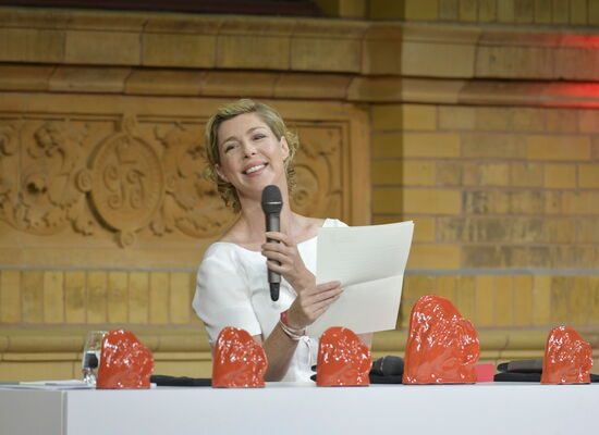 Die Schauspielerin Brigitte Zeh hält eine Laudatio beim Deutschen Hörfilmpreis 2023.  Sie steht hinter einem Pult, auf dem die Trophäen, die „ADeles“, aufgereiht stehen