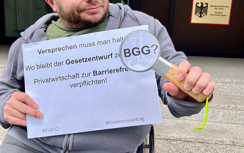 Ein Mann mit kurzen blonden Haaren hält eine Lupe und ein Schild in den Händen. Darauf steht: „Versprechen muss man halten. Wo bleibt der Gesetzentwurf zum BGG? Privatwirtschaft zur Barrierefreiheit verpflichten!“