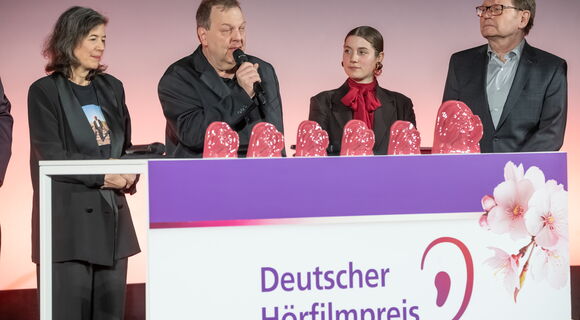 Klaus-Jörg Kaminski, blinder AD-Autor des Films „Paris, Texas“ spricht in ein Mikrofon bei der Deutschen Hörfilm-Verleihung. Neben ihm stehen sein Team und die Laudatorin des Films. Auf einem Tisch stehen sechs rosafarbene Hörfilmpreis-Trophäen.