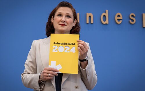 Das gelbe Heft mit dem Jahresbericht der Antidiskriminierungsstelle hält Ferda Ataman bei der Bundespressekonerenz hoch. Die unabhängigen Bundesbeauftragte hat braune, ondulierte Haare und trägt einen hellen Blazer.